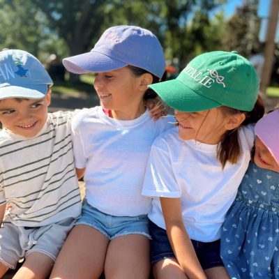 Four children sitting on a park bench wearing personalized baseball caps embroidered with their names, including 'Dean', 'Natalie' and 'Stella'.