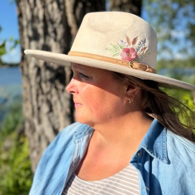 Side profile of a woman wearing a cream felt fedora embroidered with pink and blue wildflowers, standing beside a tree trunk.