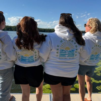 Four women standing arm-in-arm on a dock, seen from behind, wearing white hoodies printed with a 'Lake Minnetonka Social Club' sunrise-and-waves design.