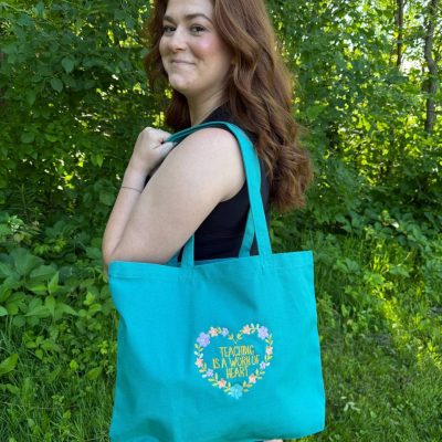 Smiling red-haired woman in a park carrying a teal canvas tote bag embroidered with a floral heart wreath and the phrase 'Teaching is a Work of Heart'.