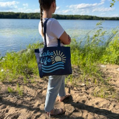 Woman standing on a sandy lakeshore carrying a navy canvas tote bag embroidered with 'Lake Life' and a sunrise-over-waves design.