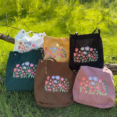 Collection of corduroy tote bags in white, yellow, black, forest green, brown and pink, each embroidered with a row of colorful wildflowers, propped on grass beside a log.