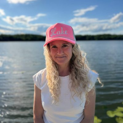 Blonde woman at a lake wearing a neon-pink foam trucker hat with 'Lake' embroidered in cream cursive script.