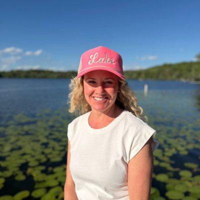 Woman wearing a pink 'Lake' trucker hat and white tee, standing in front of a lily-pad-covered lake.