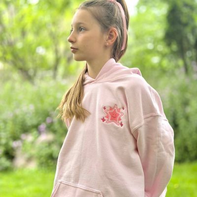 Sparkle Squad Youth Hoodie – Front Girl standing in a garden wearing a pale-pink hoodie with an embroidered pink smiling-star motif on the chest.