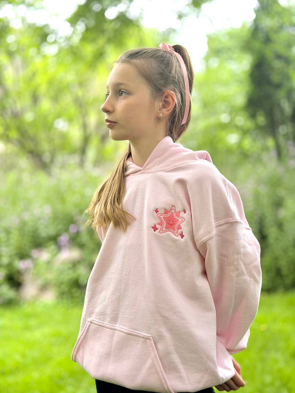 Girl standing in a garden wearing a pale-pink hoodie with an embroidered pink smiling-star motif on the chest.