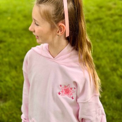 Young girl in profile outdoors wearing a light-pink hoodie with a small embroidered smiling-star design on the chest and a pink hair ribbon.