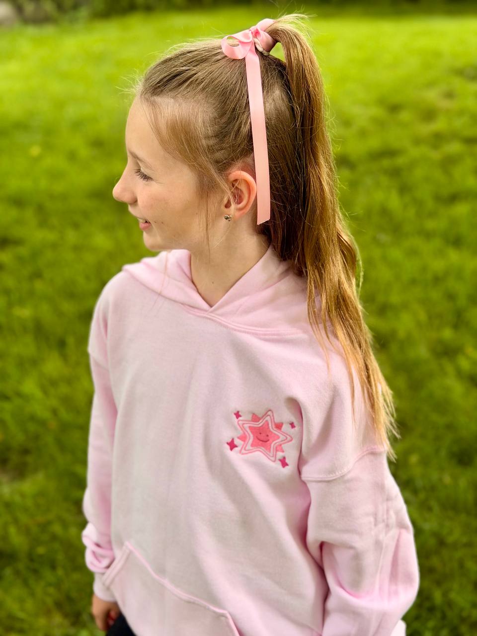 Young girl in profile outdoors wearing a light-pink hoodie with a small embroidered smiling-star design on the chest and a pink hair ribbon.