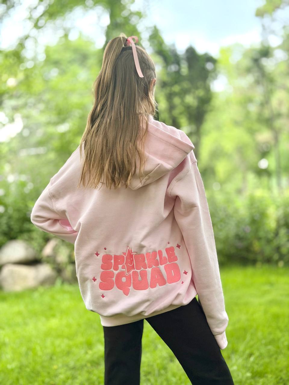 Girl seen from behind wearing a pale-pink hoodie with 'Sparkle Squad' embroidered in bubbly pink lettering and stars on the back.