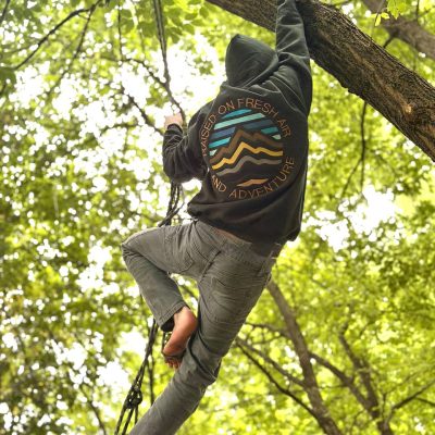 Child climbing a tree, seen from behind, wearing a dark-green hoodie with a circular 'Raised on Fresh Air and Adventure' mountain design on the back.