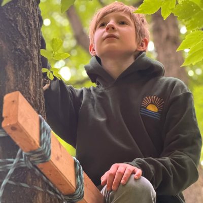 Red-haired boy sitting in a tree wearing a dark-green hoodie with a small embroidered sunrise-over-waves design on the chest.