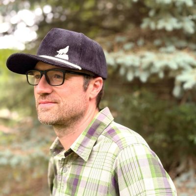 Man in glasses, side profile, wearing a navy corduroy cap embroidered with a white howling-wolf silhouette.