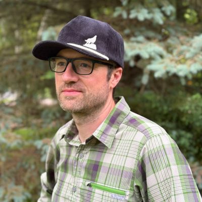 Adult Wolf Hat – Front Man wearing a dark corduroy cap embroidered with a white howling wolf, in a green plaid shirt outdoors.