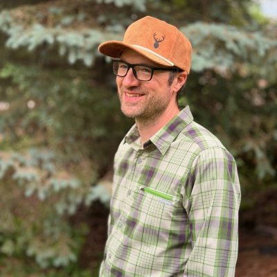 Bearded man in glasses wearing a rust-brown corduroy cap embroidered with a dark elk-head silhouette, standing in front of an evergreen tree.