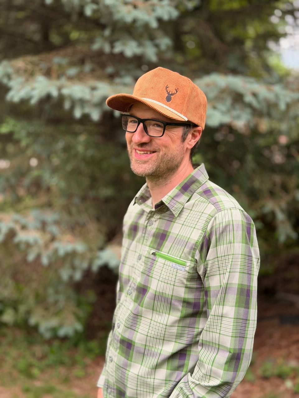 Bearded man in glasses wearing a rust-brown corduroy cap embroidered with a dark elk-head silhouette, standing in front of an evergreen tree.