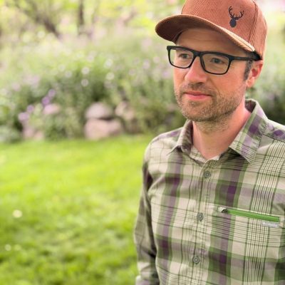 Man in glasses and a green plaid shirt wearing a brown corduroy cap embroidered with a dark elk-head silhouette.
