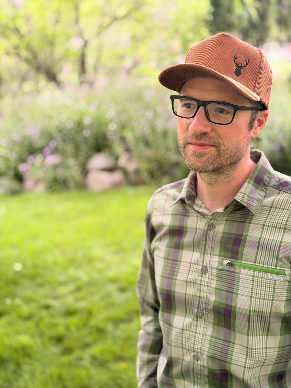 Man in glasses and a green plaid shirt wearing a brown corduroy cap embroidered with a dark elk-head silhouette.