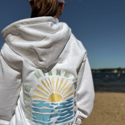 Lake Minnetonka Social Club Embroidered Sweatshirt Woman on a sandy beach seen from behind wearing a white hoodie with a colorful 'Lake Minnetonka Social Club' sunrise-over-water design on the back.