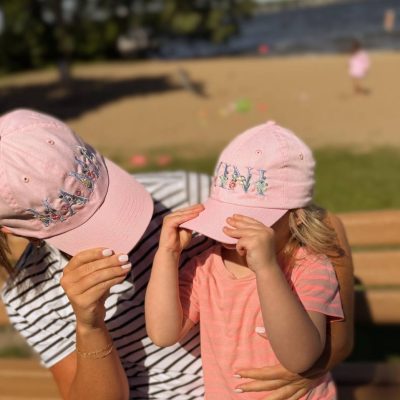 Mother and young daughter sitting by a beach holding up matching pink baseball caps embroidered 'Mama' and 'Mini' in floral lettering.