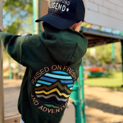 Child seen from behind on playground equipment wearing a navy 'Playground Legend' baseball cap with a forest-green hoodie.