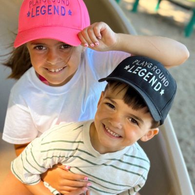 Young girl in a neon-pink cap and boy in a navy cap, both embroidered 'Playground Legend' with stars, sitting together on a playground slide.