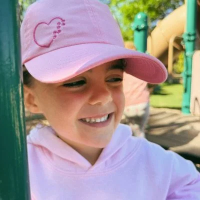 Young child at a playground wearing a soft pink Puppy Love kids cap with a heart and paw print design machine embroidered on the front