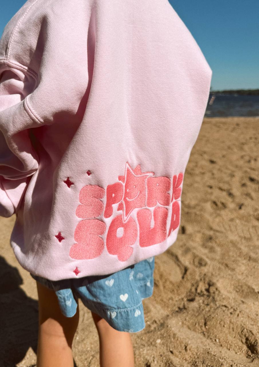 Back-view close-up of a pale-pink hoodie embroidered with 'Sparkle Squad' in bubbly pink letters and a smiling star, worn by a child at the beach.