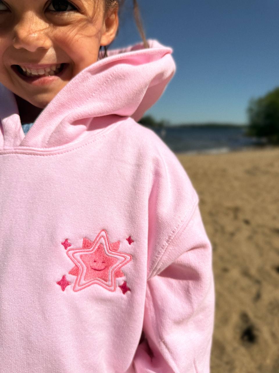 Close-up of a smiling girl on a beach wearing a pale-pink hoodie with an embroidered pink smiling-star design on the chest.